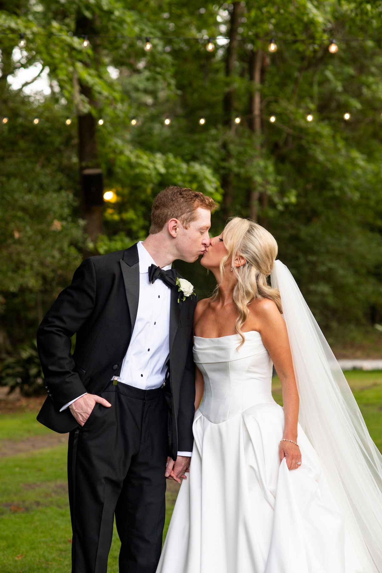 A bride and groom share a romantic kiss outdoors, she in a white strapless gown with veil, he in a black tuxedo.