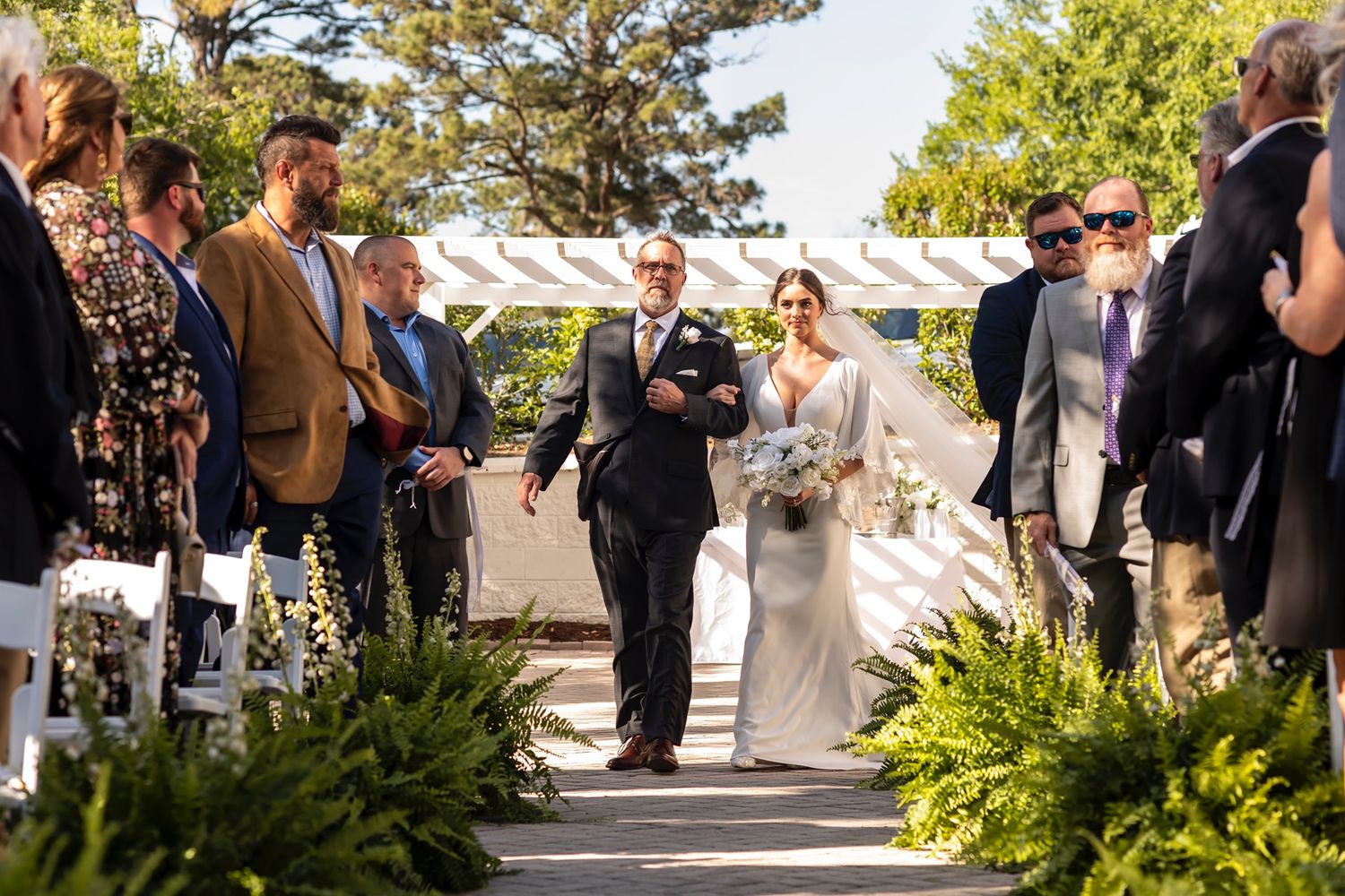 A couple walks down an outdoor ceremony aisle lined with ferns and guests during a sunny wedding ceremony.
