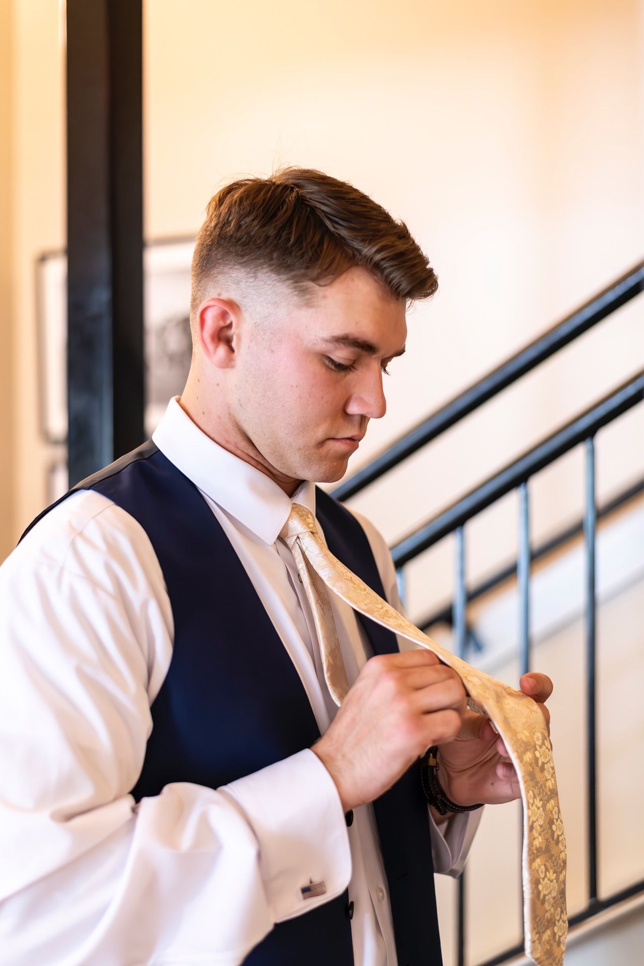 Man in white dress shirt and navy vest adjusts gold tie while standing near stairway railing.