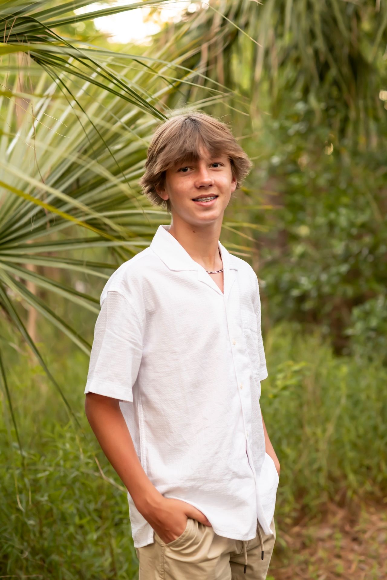 Smiling teenage boy in white linen shirt standing among tropical palm trees and lush green foliage outdoors.