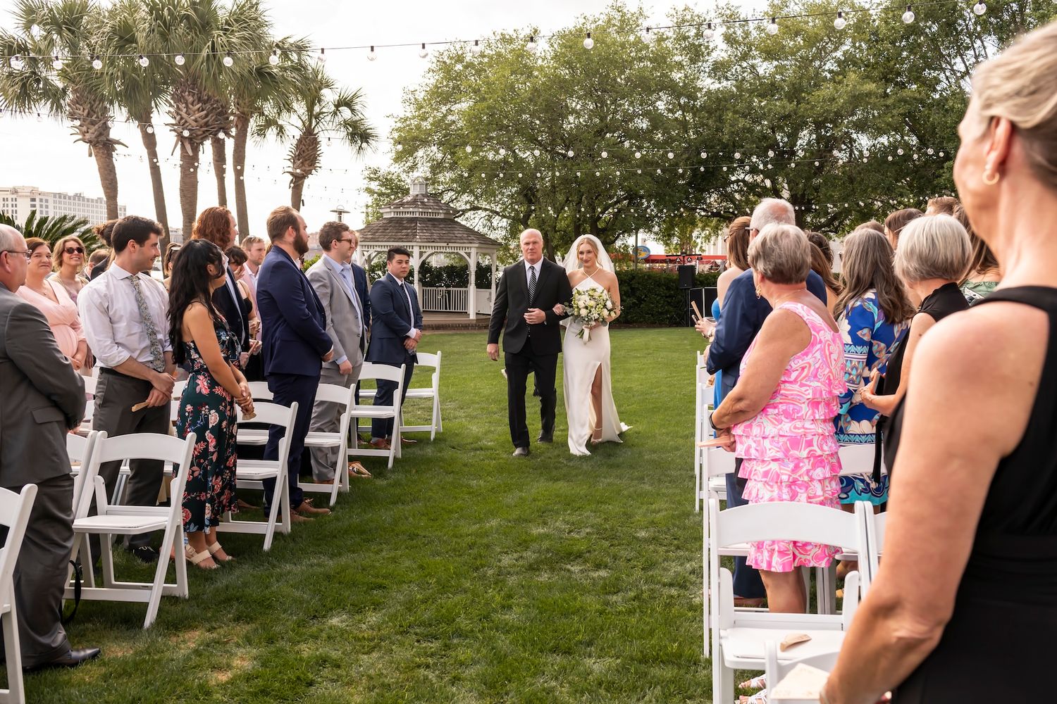 Bride walks down outdoor aisle with escort at elegant garden wedding ceremony with white chairs and palm trees.