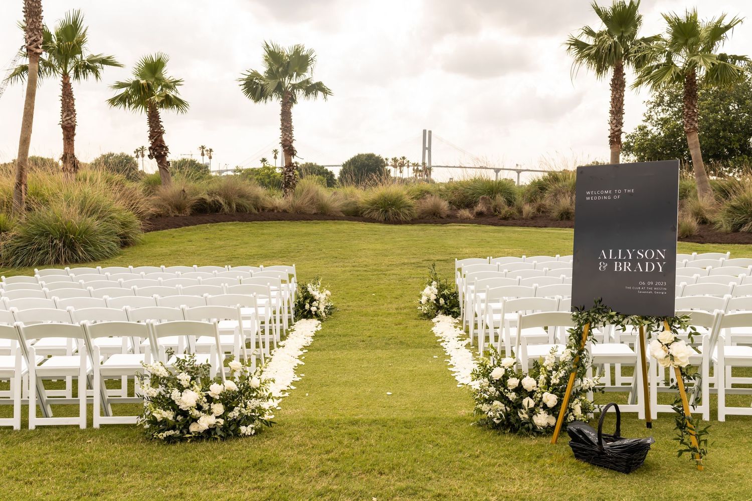 Outdoor wedding ceremony setup with white chairs, floral arrangements, and a sign reading Allyson and Brady on a green lawn.