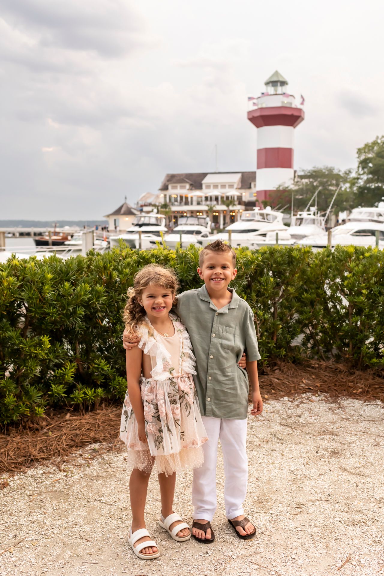 Two children walking on a shell path with boats and a lighthouse visible in the background at a marina.