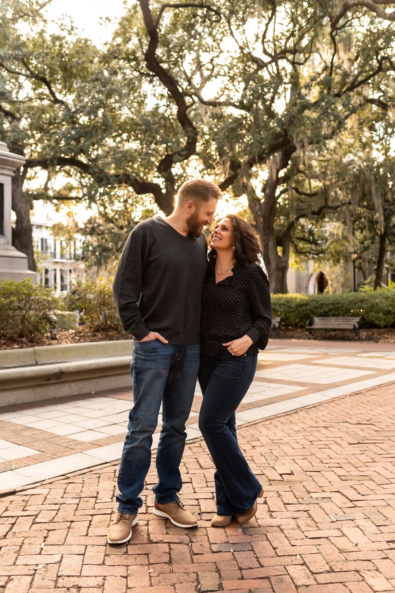 A couple shares an intimate moment on a brick path beneath Spanish moss-draped oak trees at sunset.