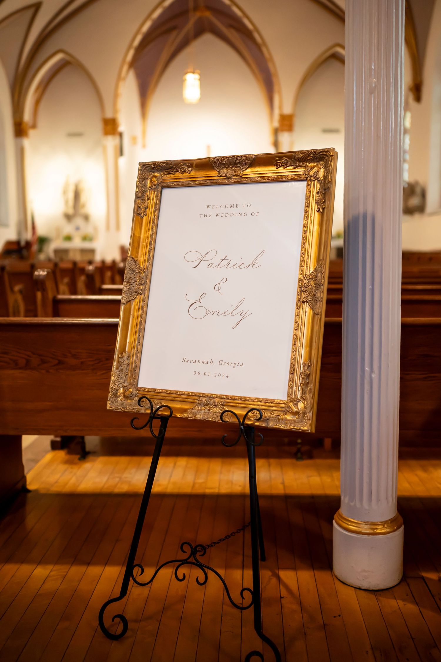 A gold ornate frame displaying a wedding welcome sign stands on an easel inside a church with vaulted arches.