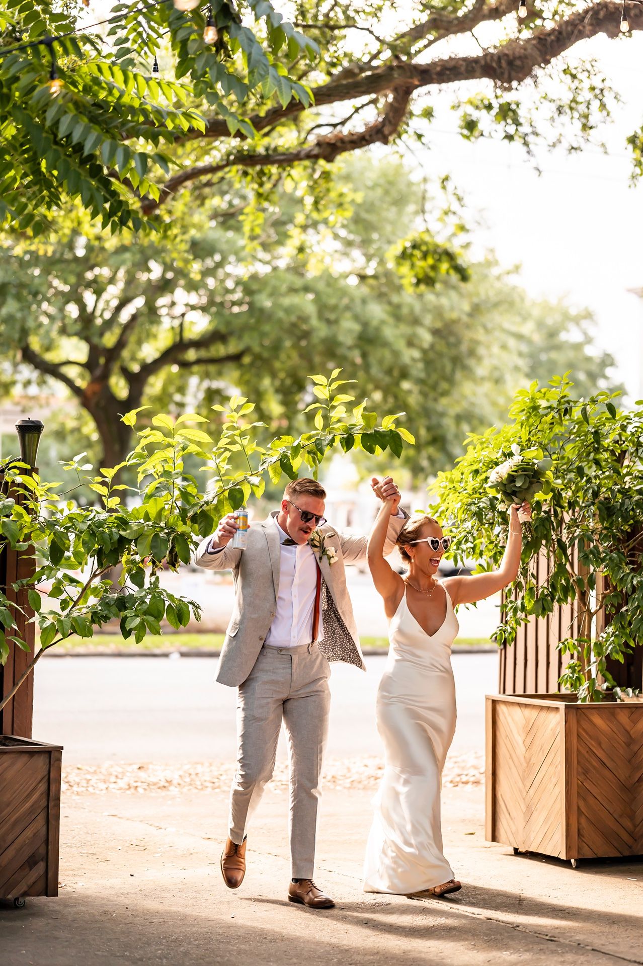 Joyful couple celebrates while walking through a garden pathway beneath lush trees and planters on a sunny day.