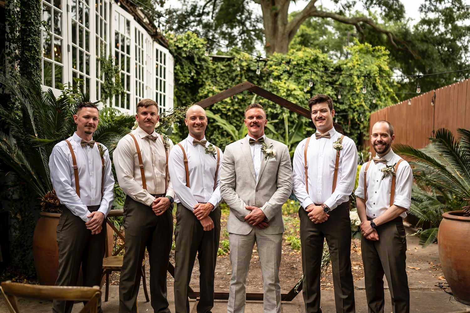 Groomsmen wearing gray suits and suspenders pose in a row outdoors at a wedding venue.
