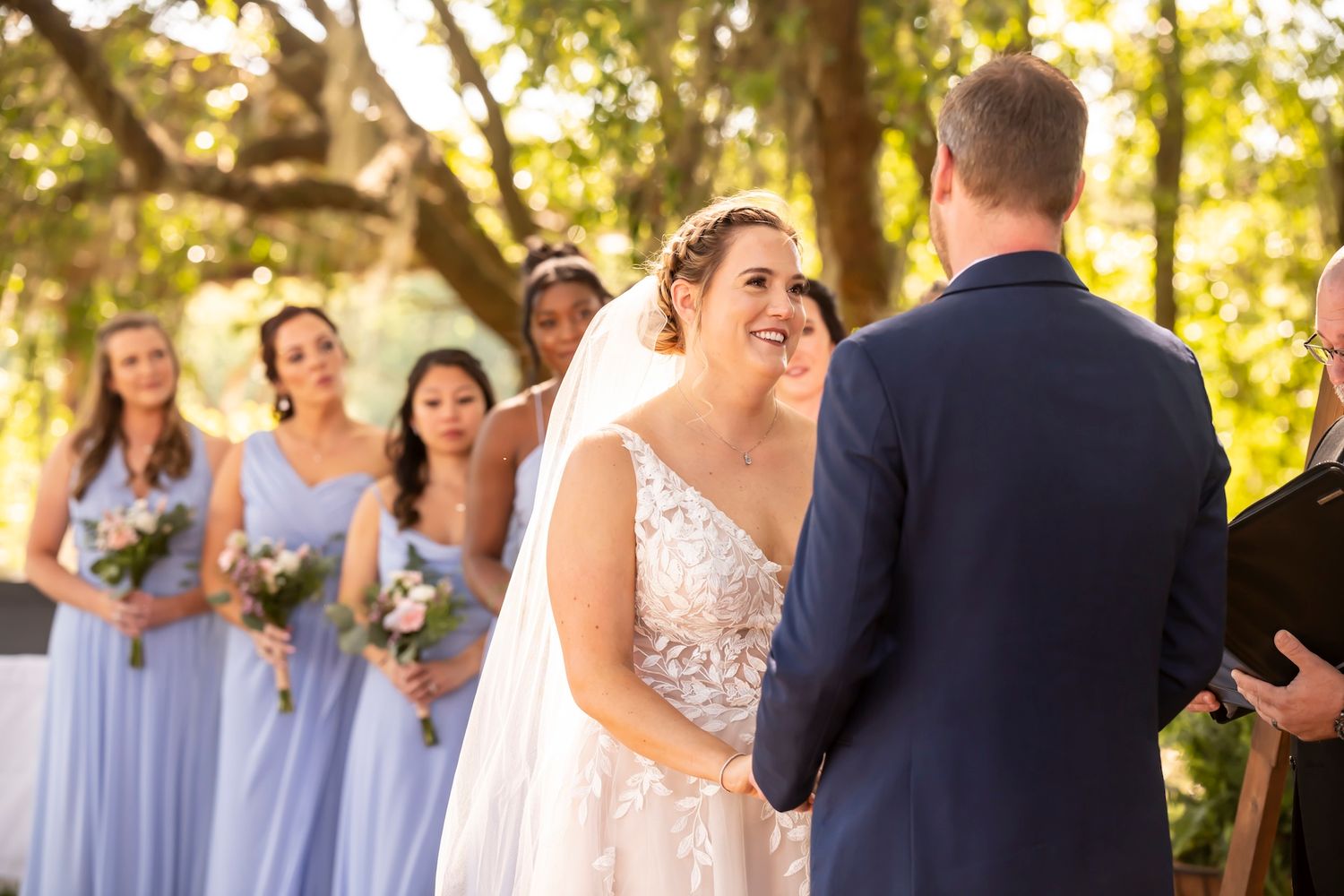 Wedding ceremony in sunlit outdoor setting with bridesmaids in light blue dresses watching the couple exchange vows.