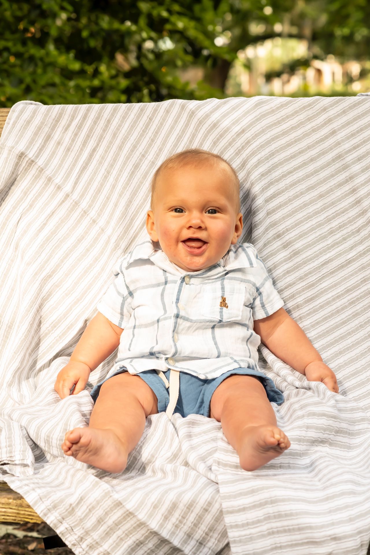 Smiling infant in blue and white outfit relaxing on striped blanket with natural outdoor backdrop.
