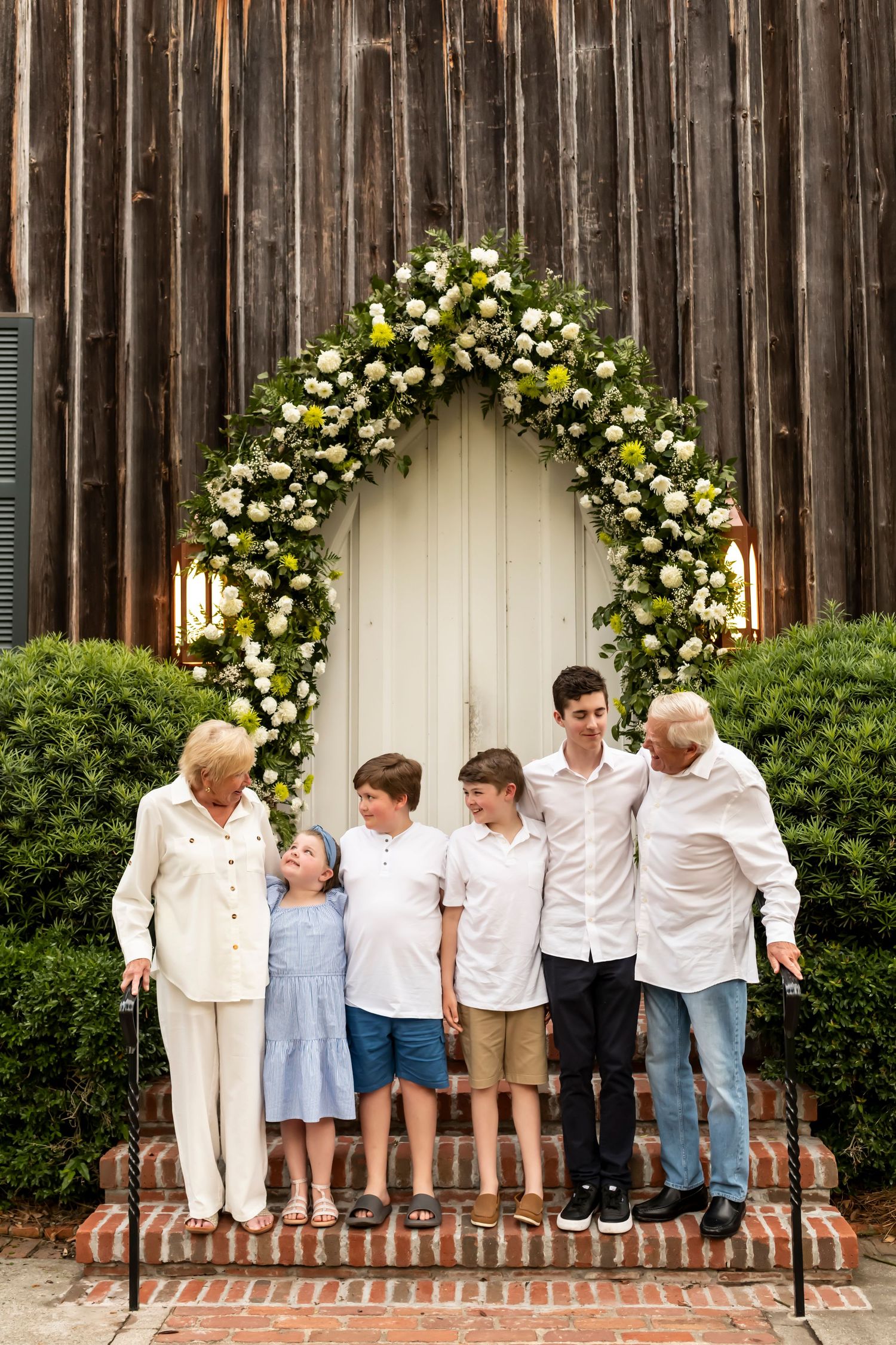 Multi-generational family standing under elegant floral arch by rustic wooden barn wall with lanterns.