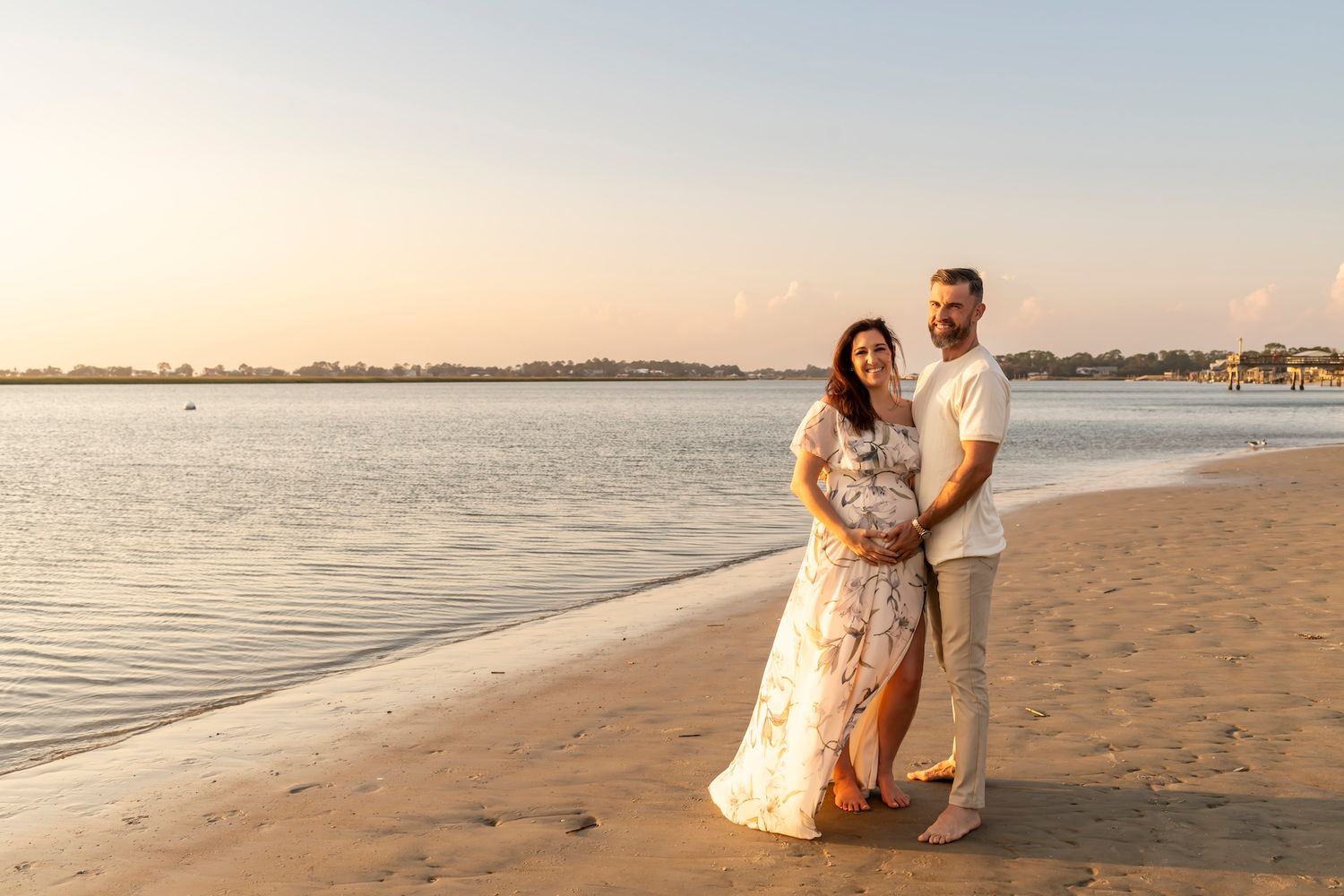 A couple embraces on a sandy beach at golden hour with calm waters and distant shoreline in view.
