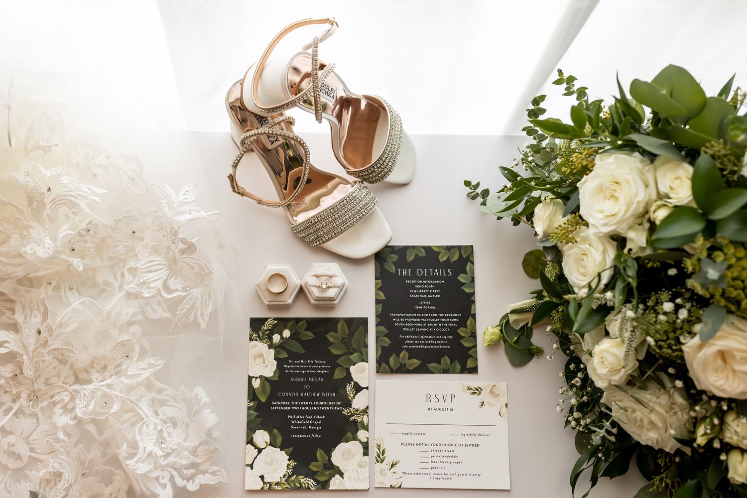 Wedding details flatlay with black floral invitations, bridal shoes, and white rose bouquet on light background.