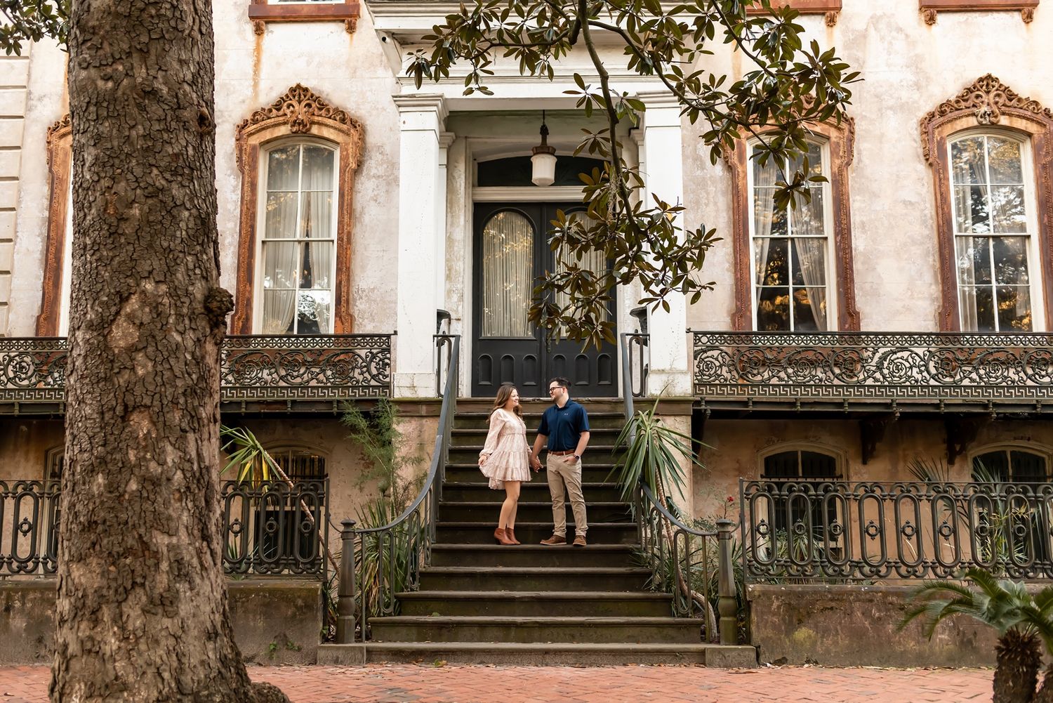 Couple posing on ornate staircase of historic Victorian-style home with lush trees.