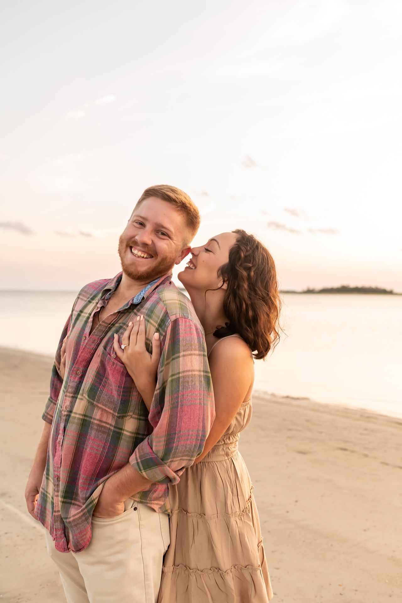 A joyful couple laughs together on a beach at sunset while dressed in casual attire.