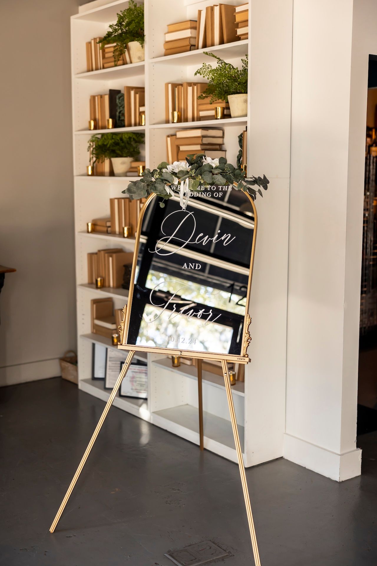 Black chalkboard wedding sign on wooden easel displayed in front of white built-in bookshelves decorated with books and plants.