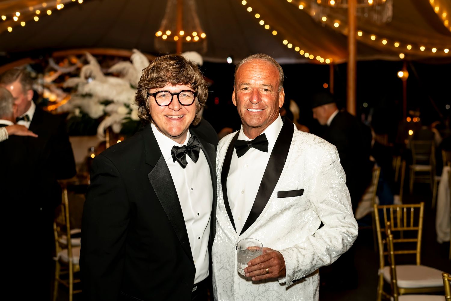 Two people in formal evening attire, one wearing glasses and a black tuxedo, the other in a white dinner jacket.