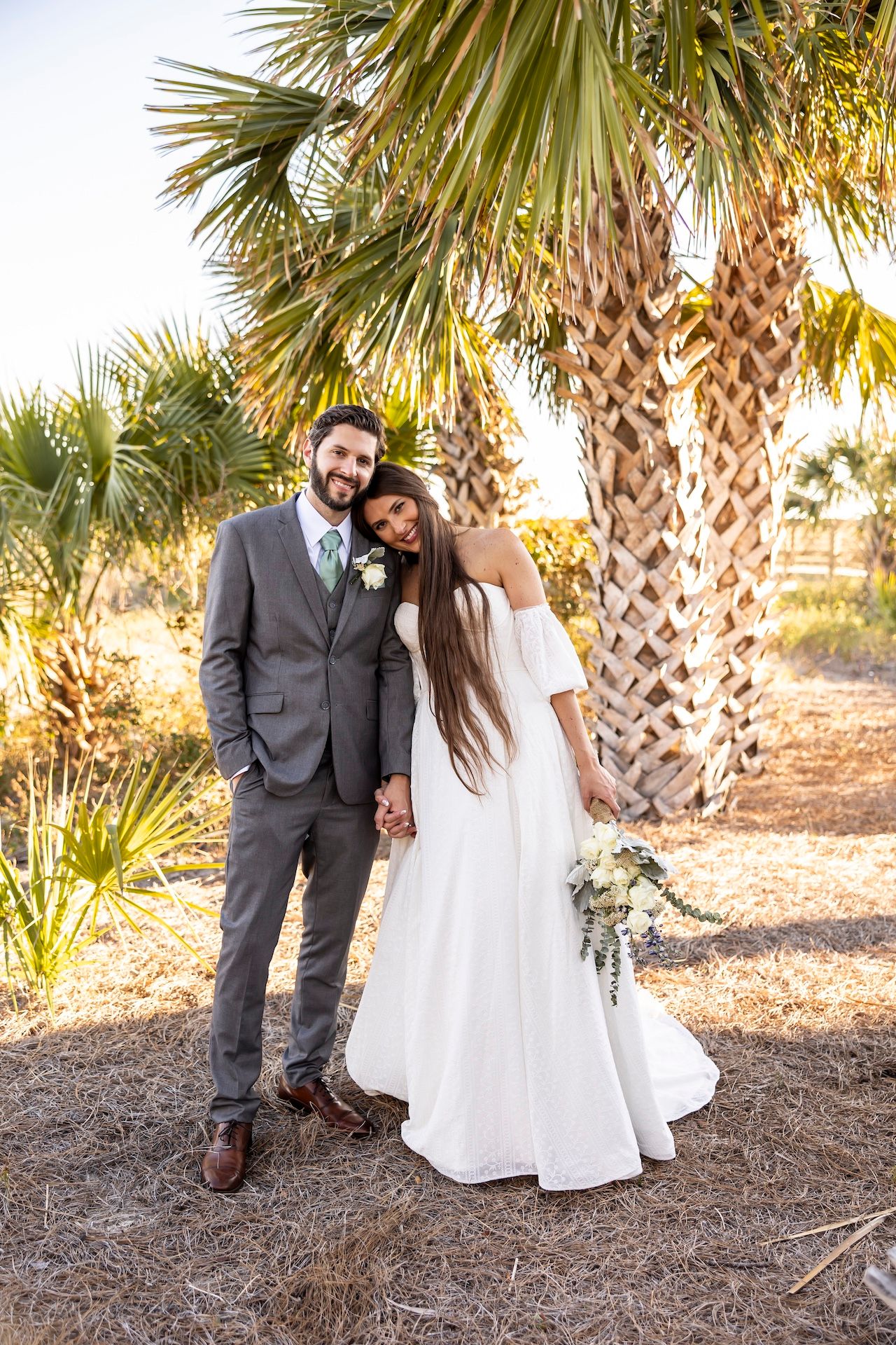 A newlywed couple poses together under palm trees at a beach wedding venue during sunset.