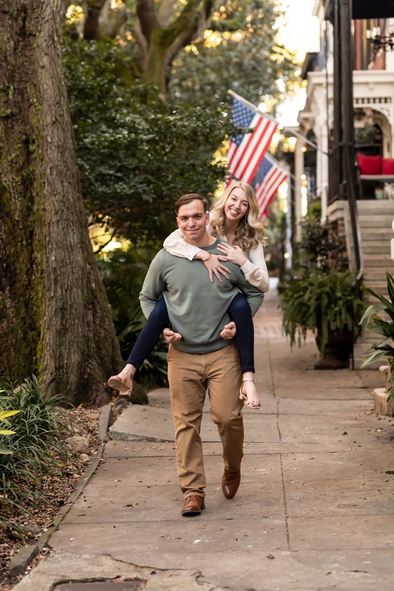 A couple shares a playful piggyback ride on a tree-lined sidewalk with an American flag visible in the background.