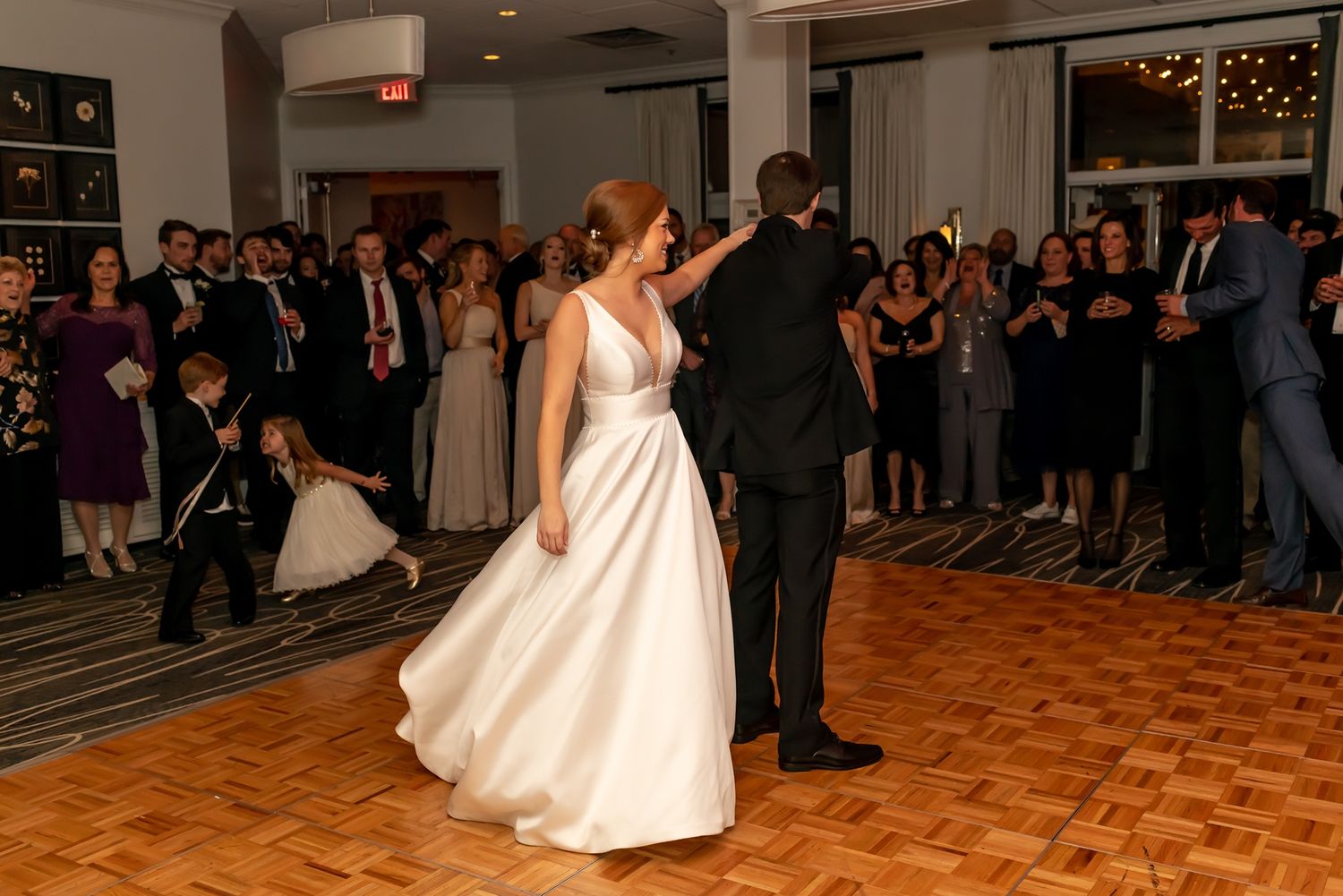Bride and groom share their first dance on a parquet floor during a wedding reception surrounded by guests.