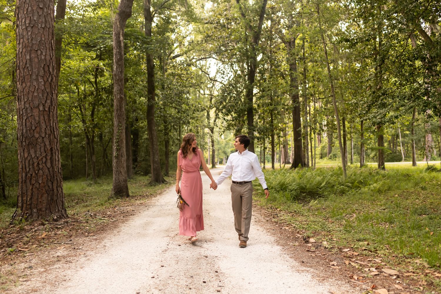 A couple walks hand in hand down a scenic forest path with tall pine trees and sunlit greenery.
