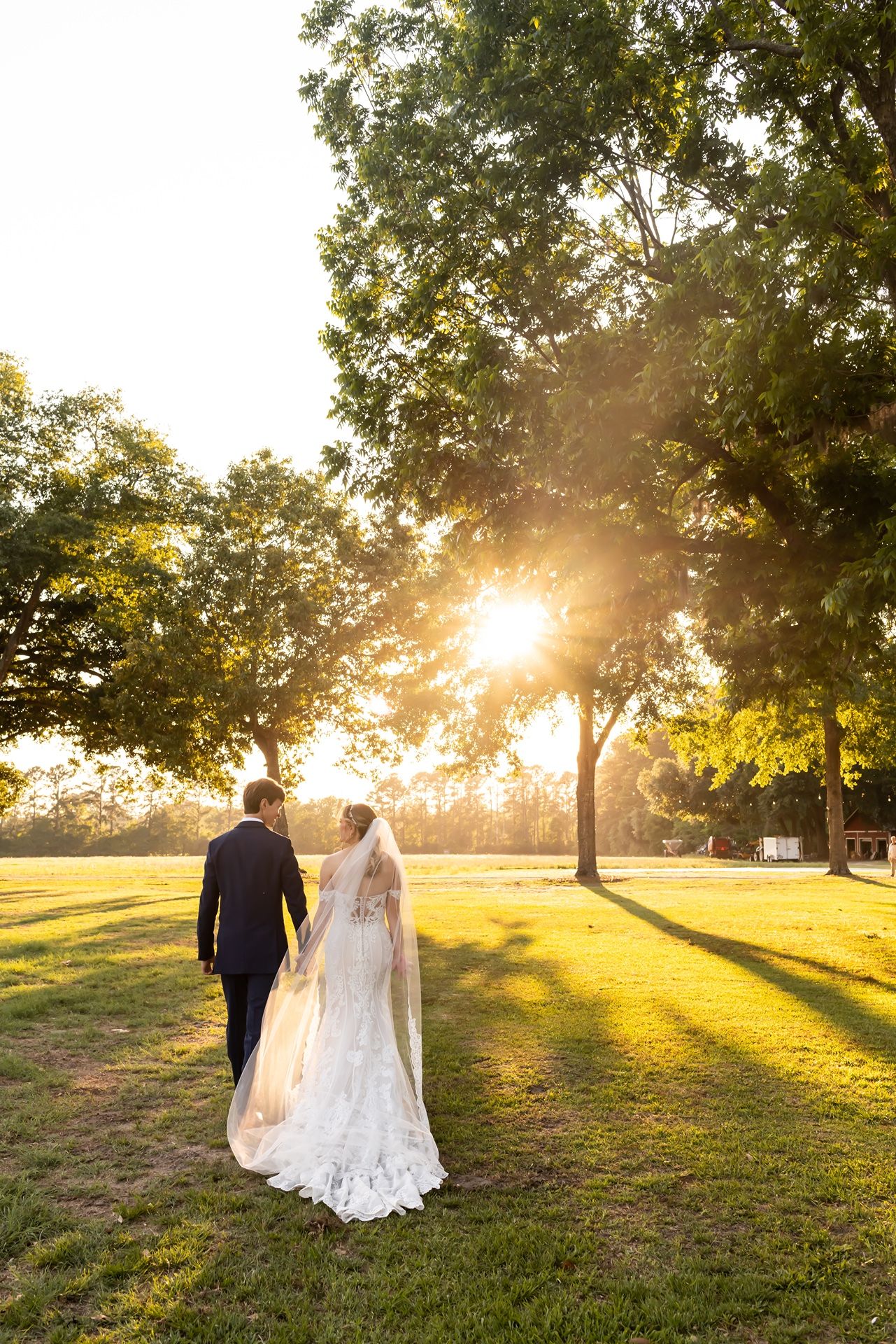 A couple in wedding attire walks across a sunlit grassy lawn with golden sunlight streaming through trees at sunset.