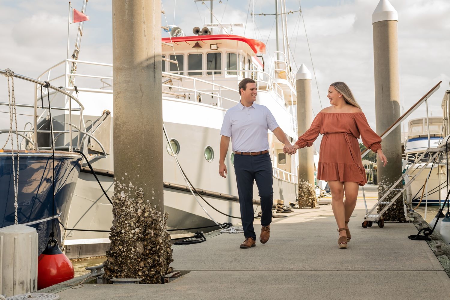 A couple holding hands walks along a marina dock with boats and a lighthouse in the background on a cloudy day.