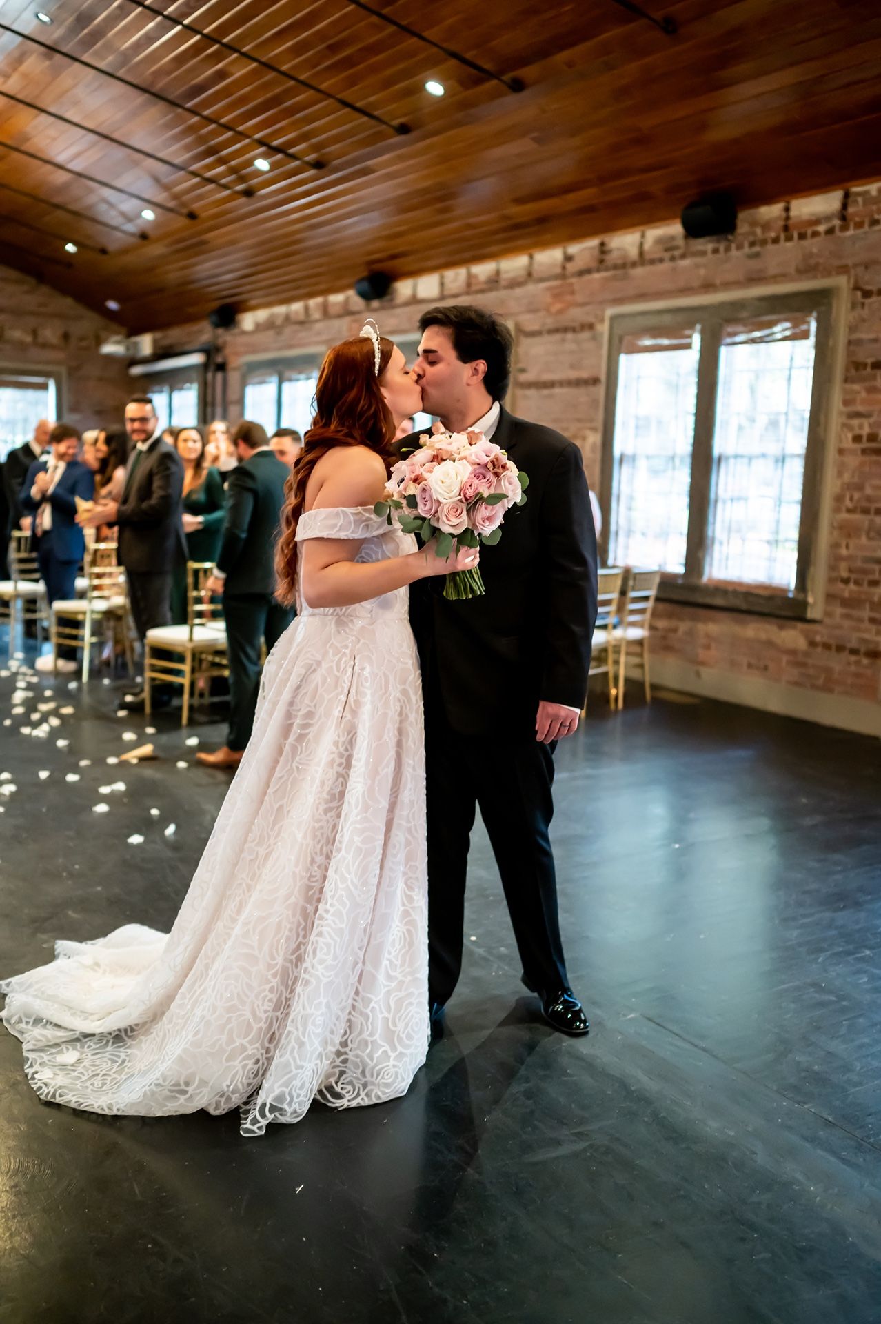 A bride and groom share an intimate first dance in an industrial-style venue with exposed brick walls and wooden beams.