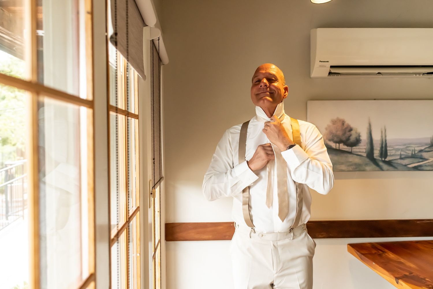 A person in a crisp white suit adjusts their tie while standing near sunlit windows in an elegant interior space.