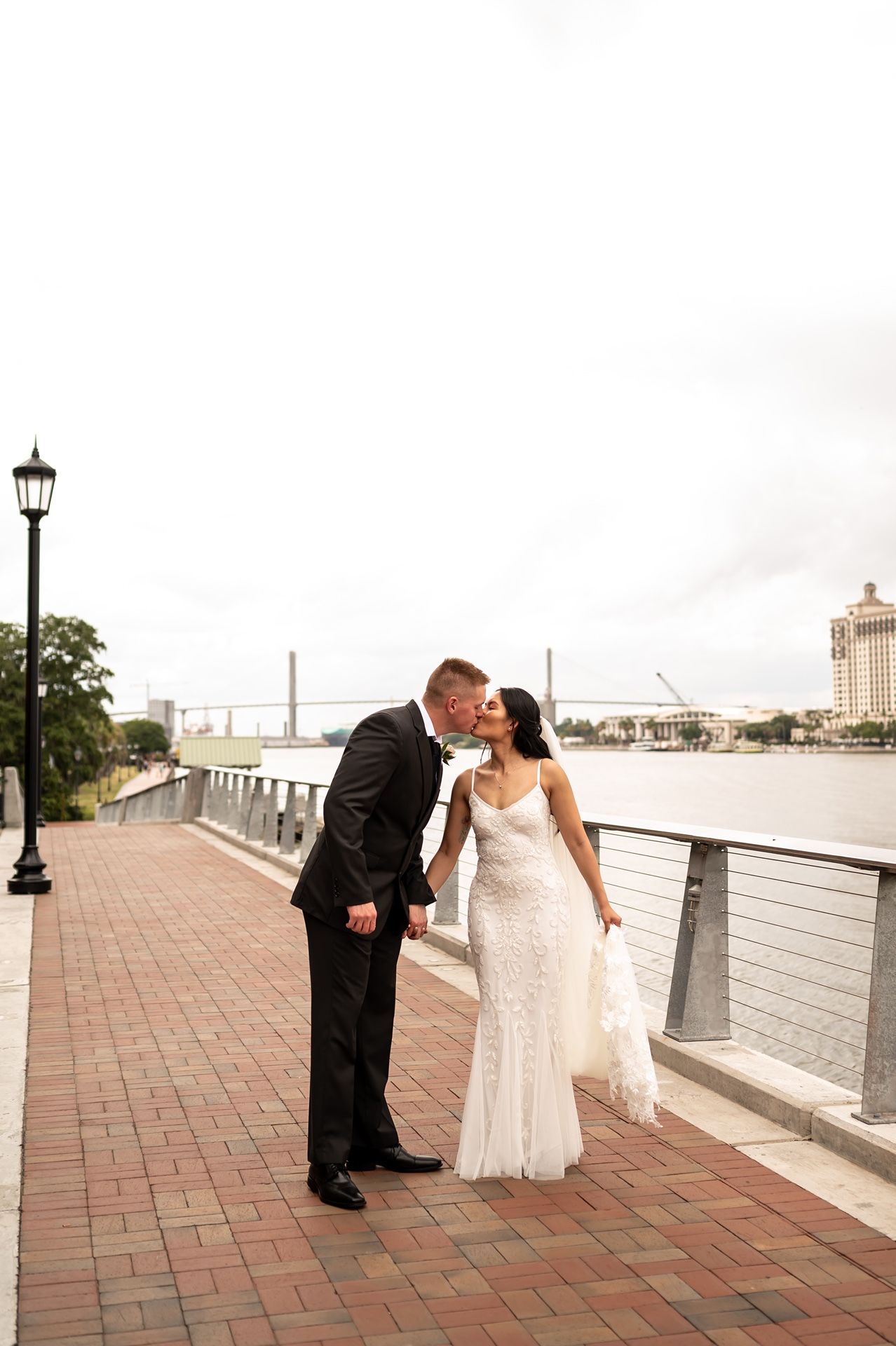Newlywed couple shares a romantic moment on a brick waterfront promenade with decorative lamp post and river view.