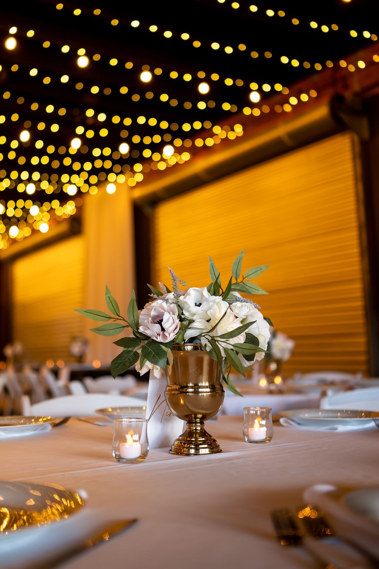 Elegant wedding table centerpiece with white flowers in gold vase under warm string lights with dreamy bokeh effect.
