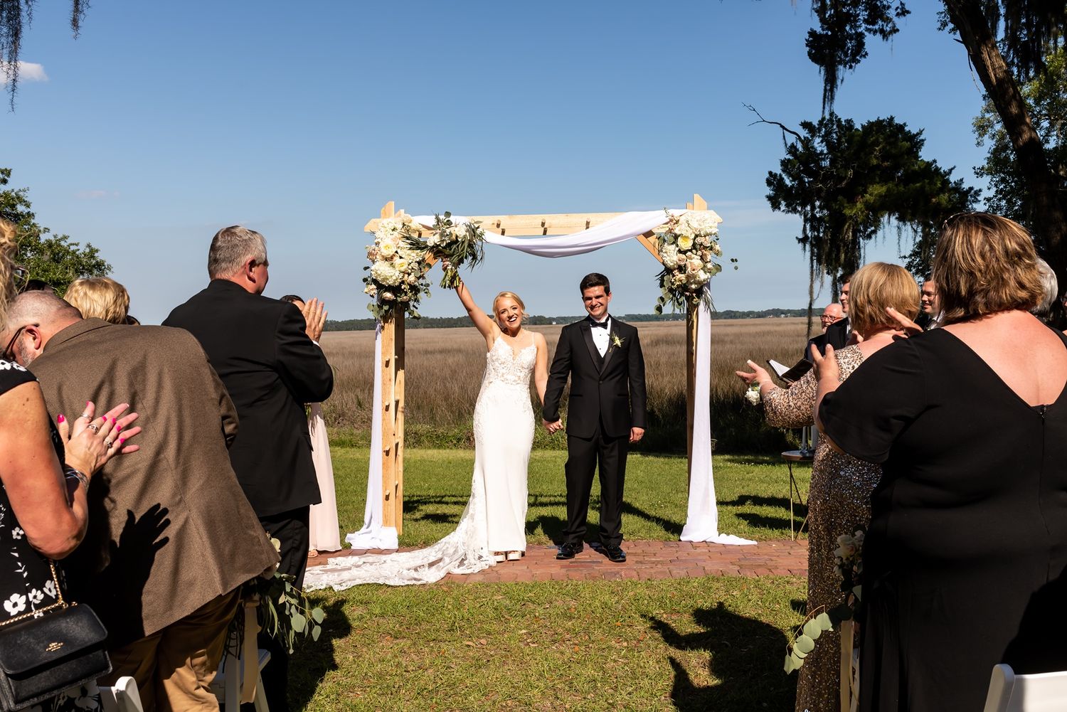 Panoramic view of an outdoor wedding ceremony with guests seated on chairs and a decorated arch against a scenic backdrop.