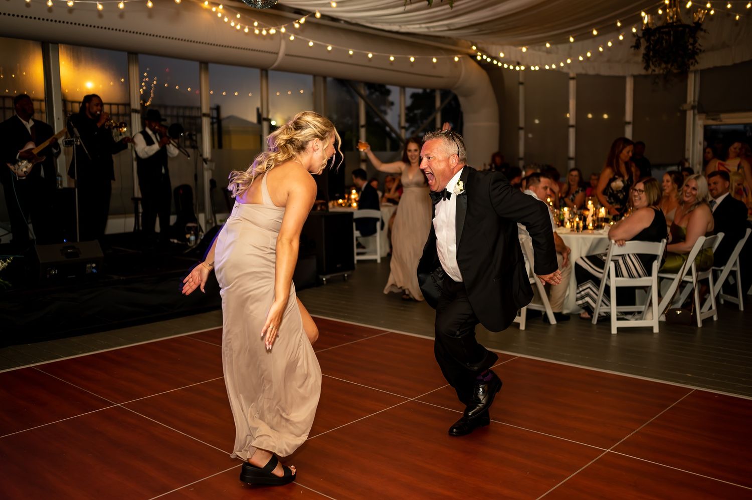 Two people dancing energetically on a wooden dance floor at an evening reception under string lights and surrounded by guests.