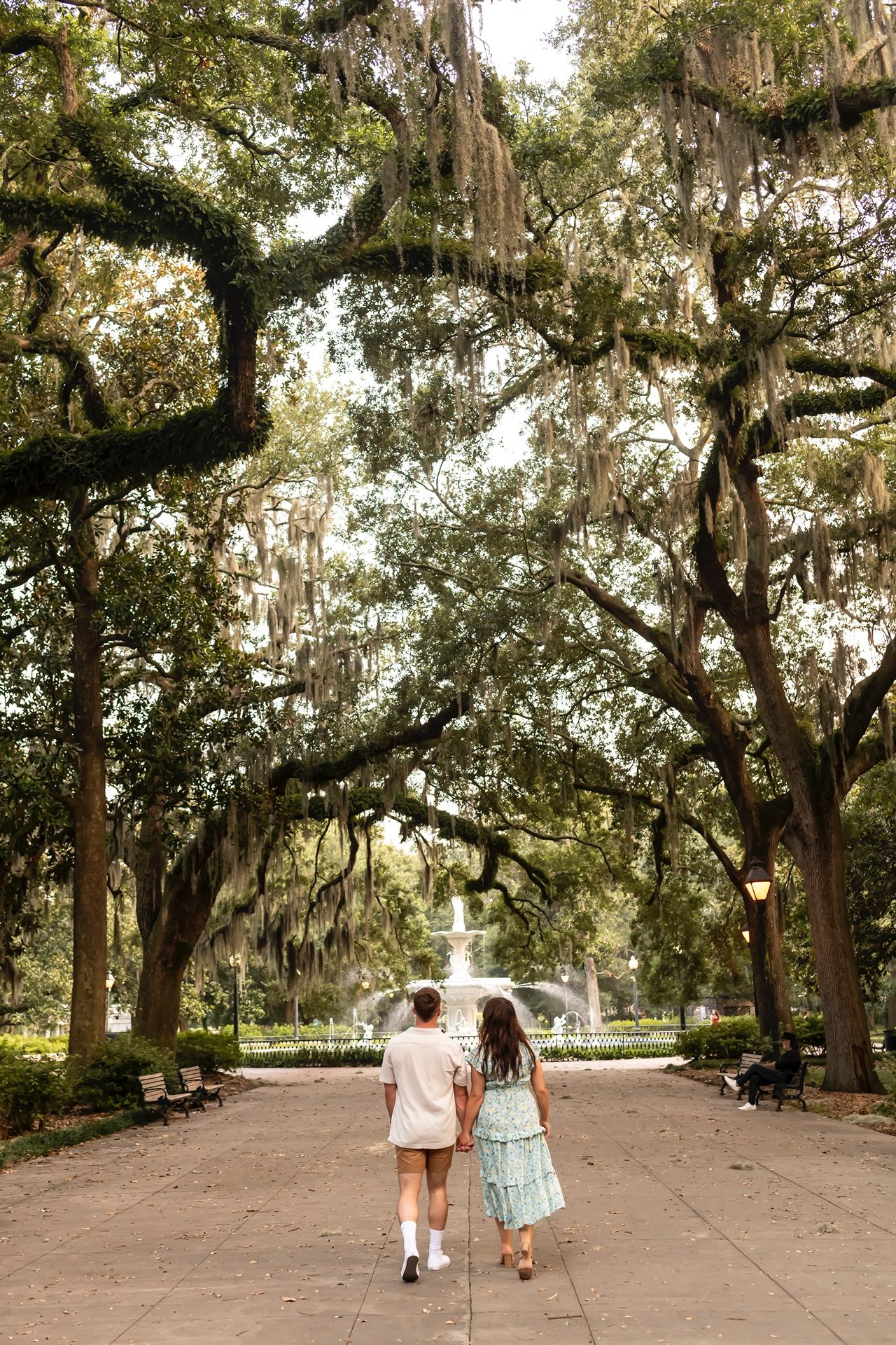 A couple strolls down a tree-lined path with Spanish moss hanging from live oaks and a fountain visible in the distance.