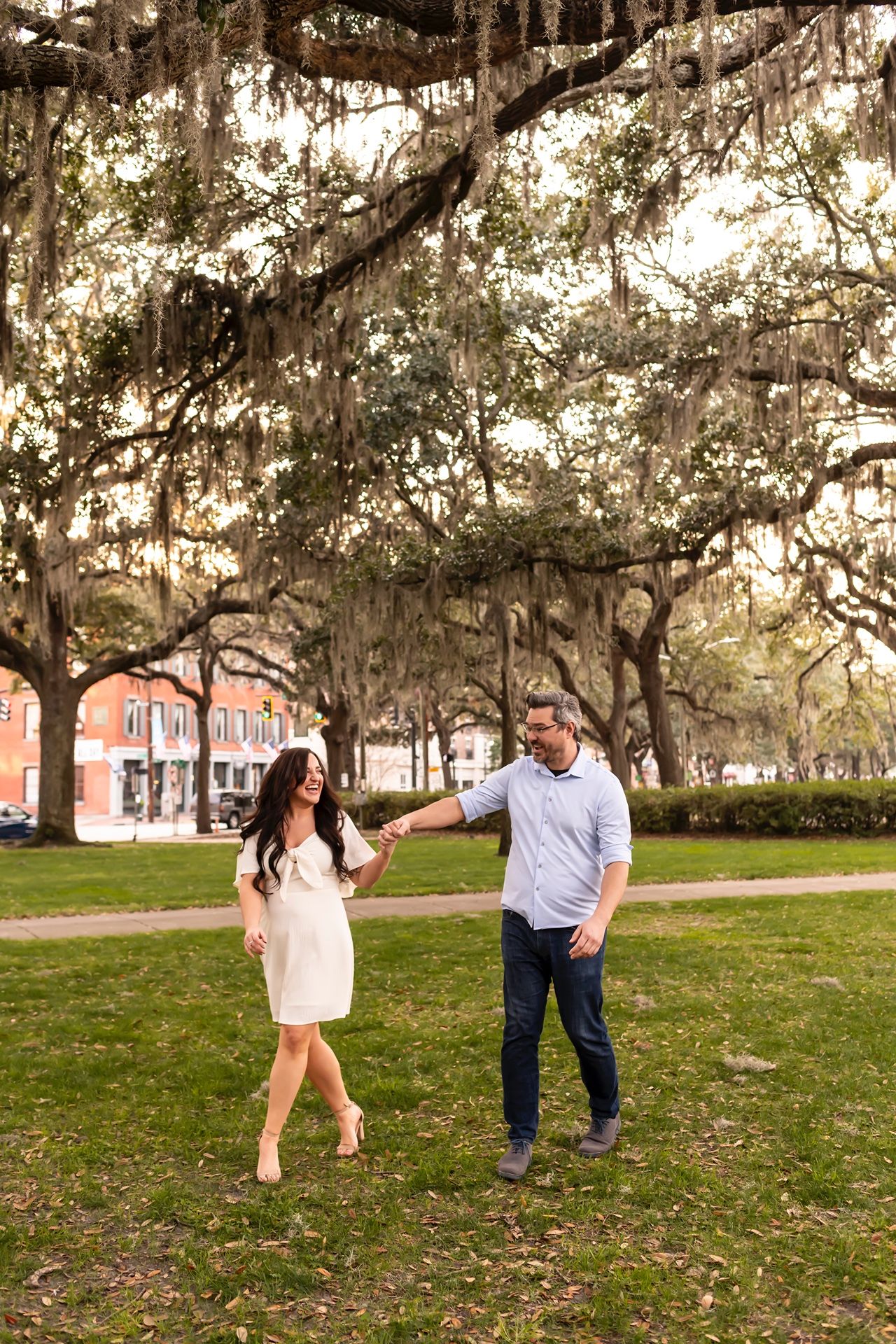 A couple walks under Spanish moss-draped oak trees at sunset in a scenic Southern park setting.