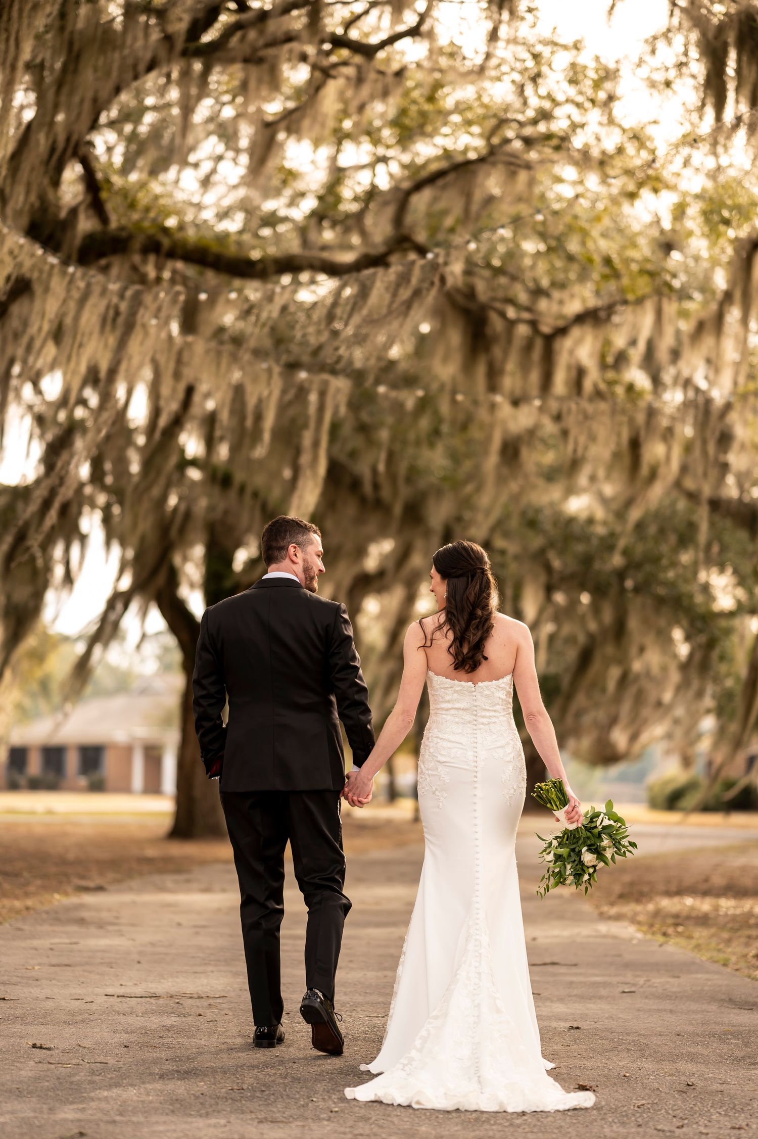 A couple in wedding attire walks along a scenic path beneath Spanish moss-draped oak trees at sunset.