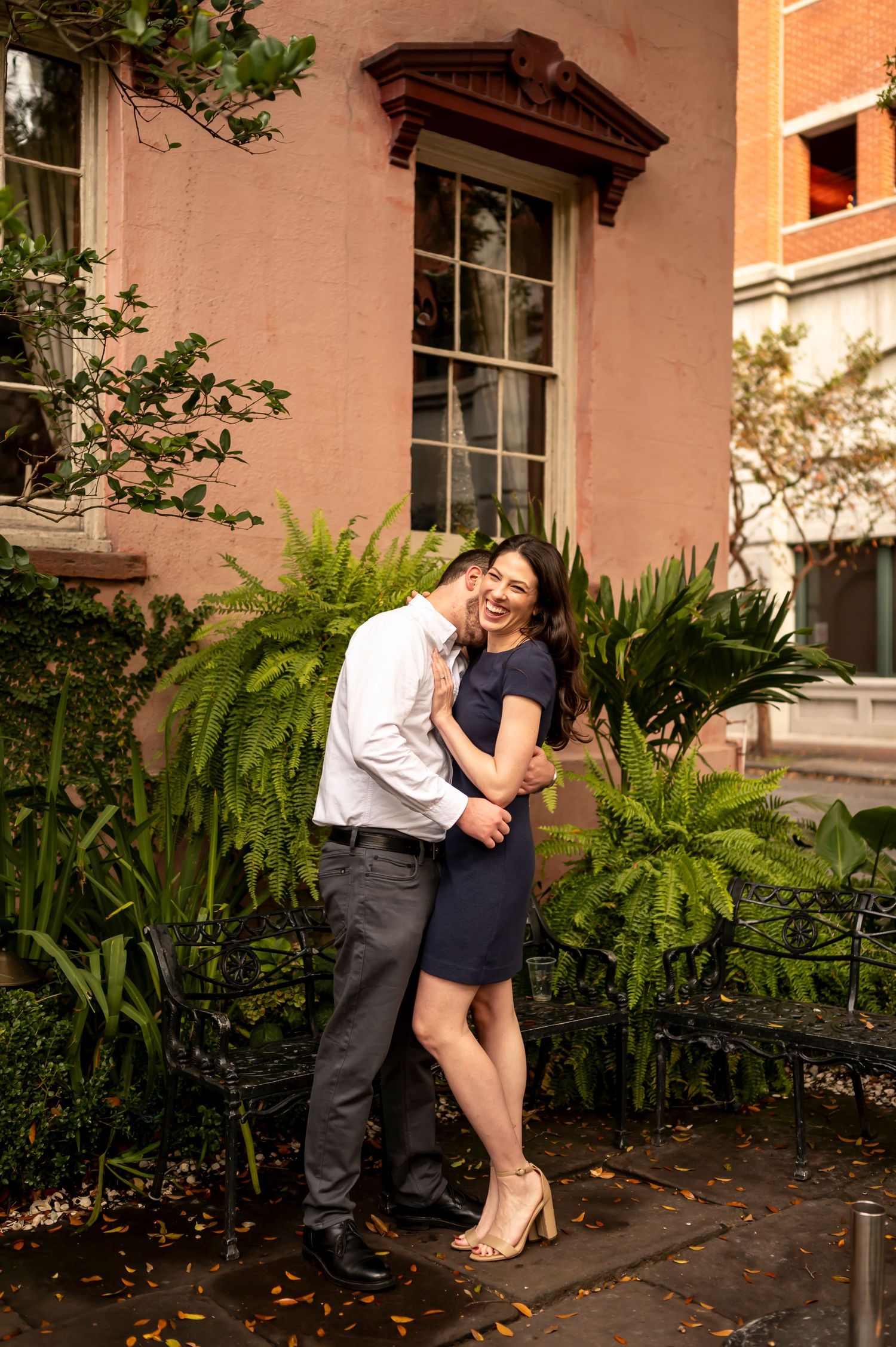 A couple embraces in front of a pink building with ornate window trim and lush ferns in an outdoor garden setting.