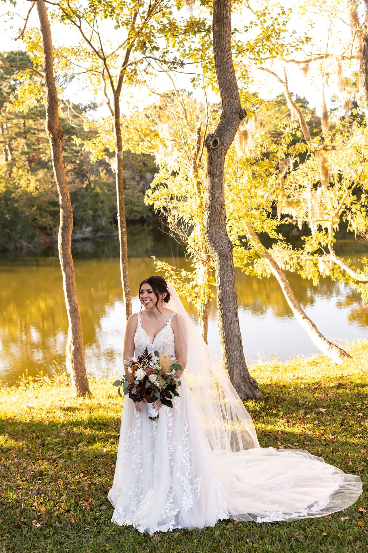 A bride in a flowing white lace dress with long train poses by a serene lake at sunset among tall trees.