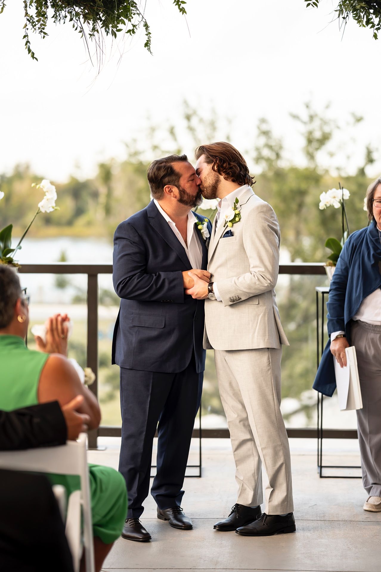 Two people in suits share a tender moment during a wedding ceremony while guests look on at an outdoor venue.