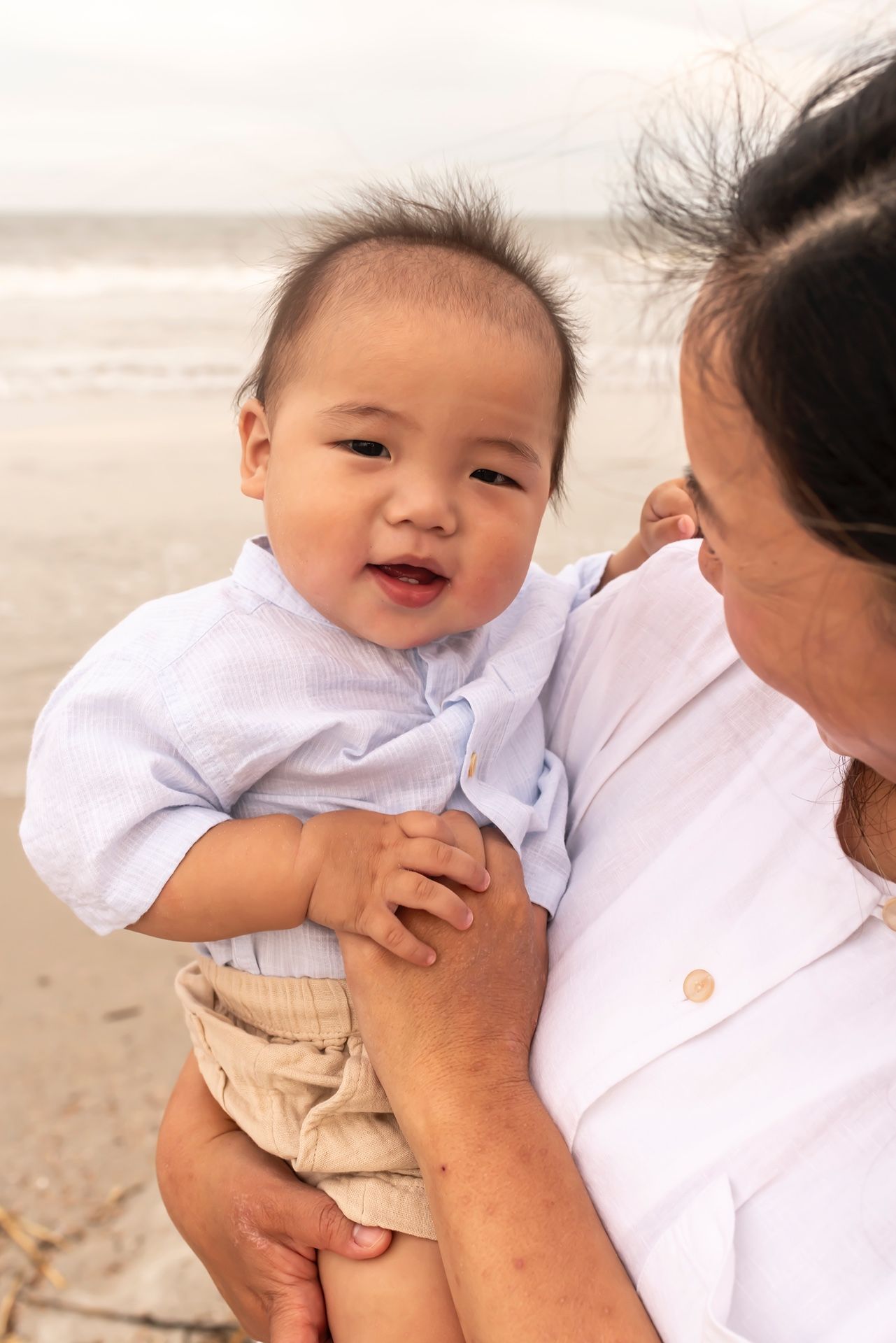 A laughing baby in white clothing is held by parent on sunny beach with ocean waves in background.