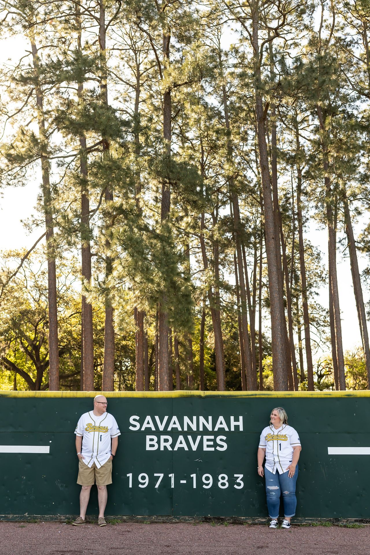 Two people stand in front of a dark green wall with 'Savannah Braves 1971-1983' text in a baseball stadium with tall pine trees.