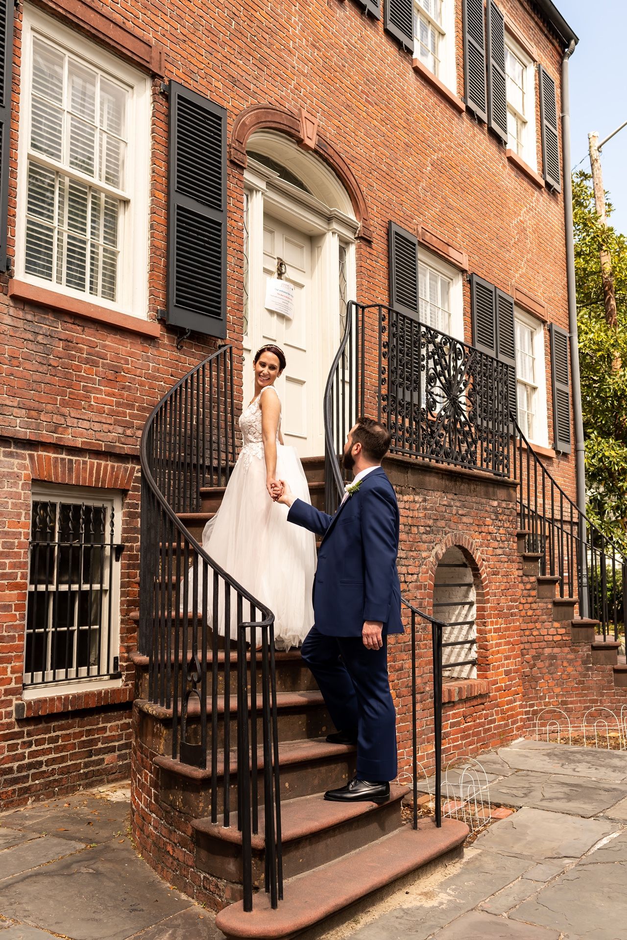 A couple in wedding attire stands on the front steps of a historic red brick townhouse with black shutters and railings.