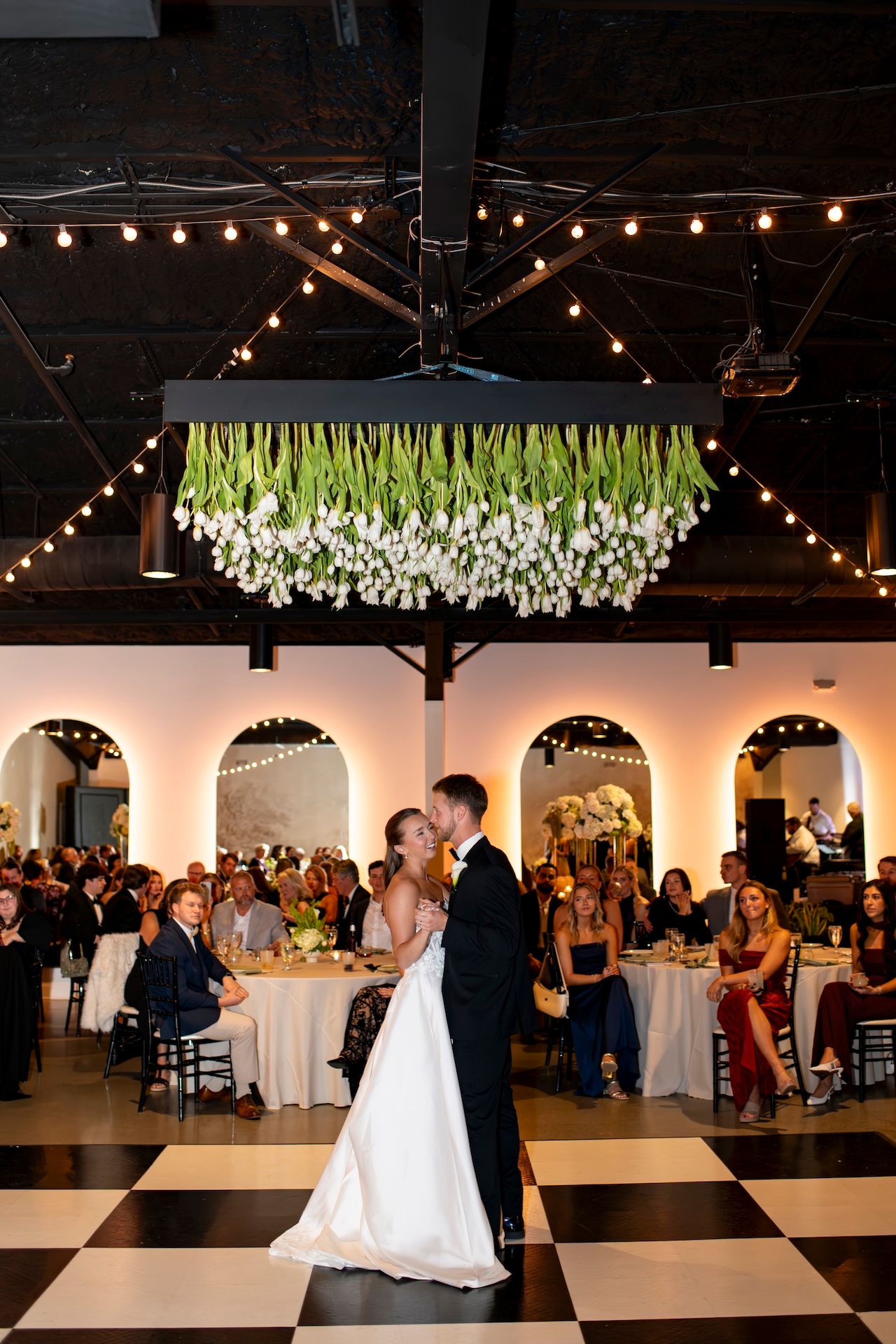 Bride and groom share first dance on checkered floor beneath hanging white tulips at The Clyde in Savannah, Georgia.