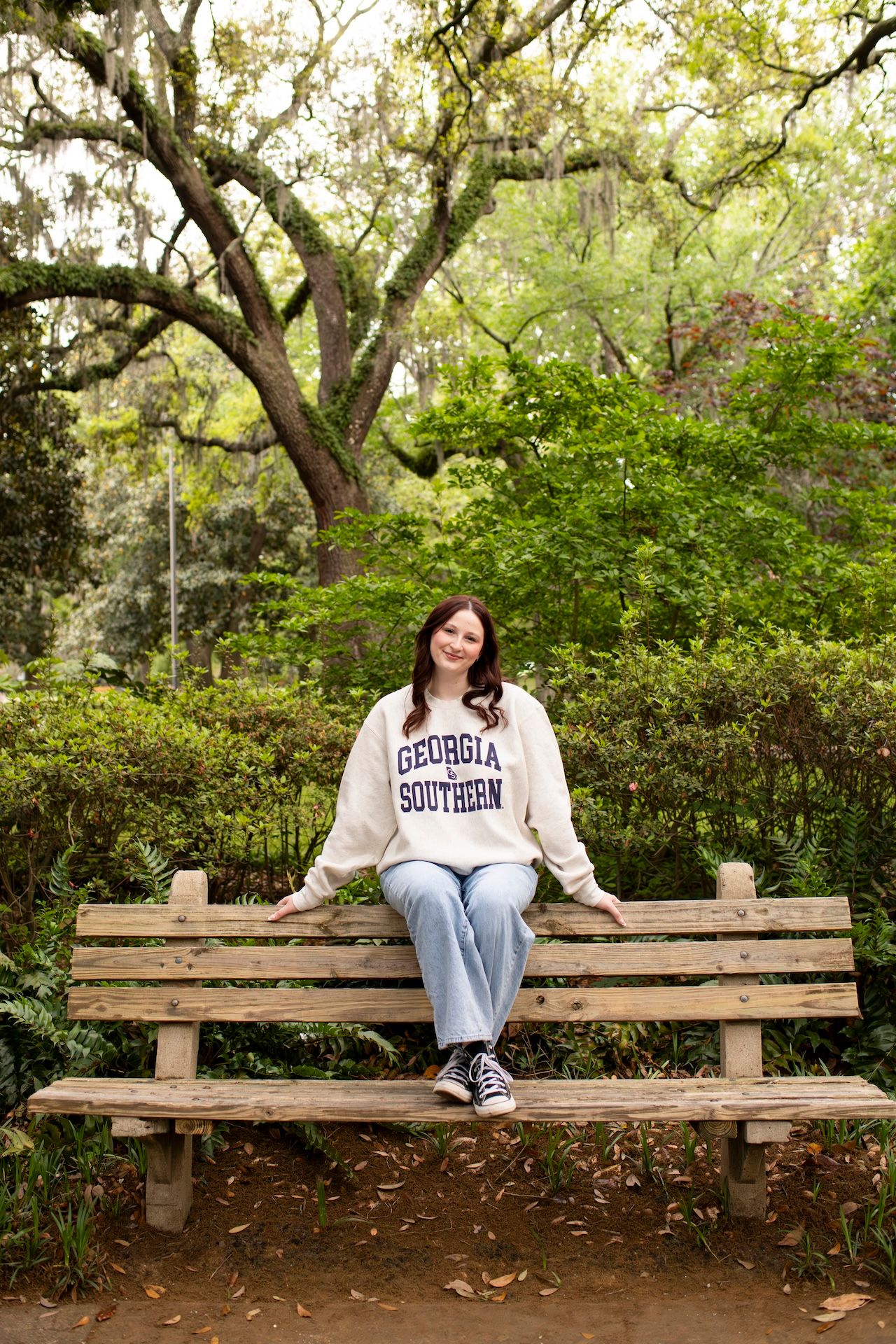 Senior in Georgia Southern sweatshirt sitting on wooden park bench surrounded by lush green trees in Forsyth Park.