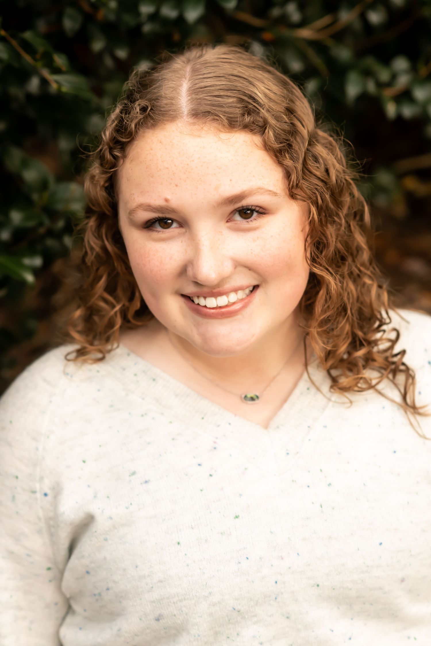 Senior portrait of a person in white sweater with curly hair against natural foliage background.
