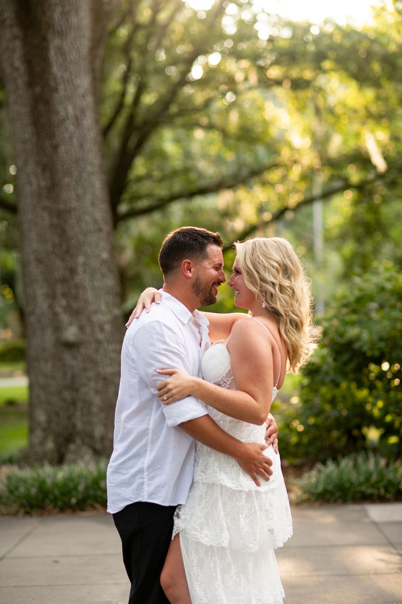 A couple shares a romantic kiss in Forsyth Park with tall trees creating a natural backdrop.