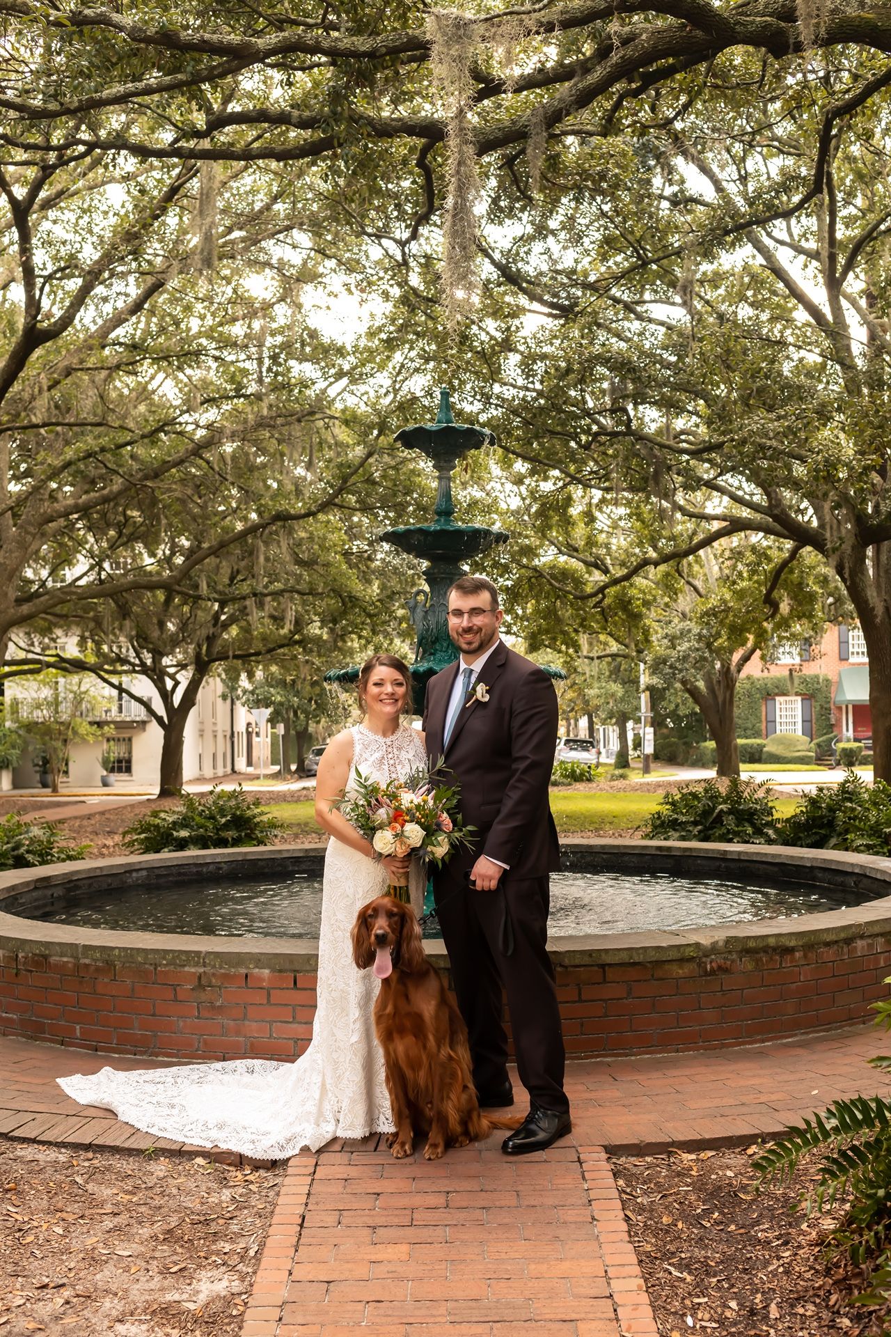 Wedding couple stands with their golden retriever by a brick fountain in a tree-lined park setting at dusk.
