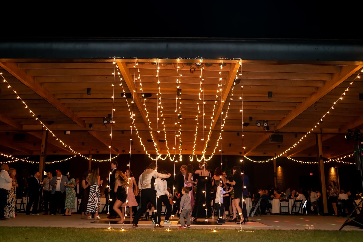 String lights illuminate an outdoor pavilion at night where people dance and gather on a lawn.