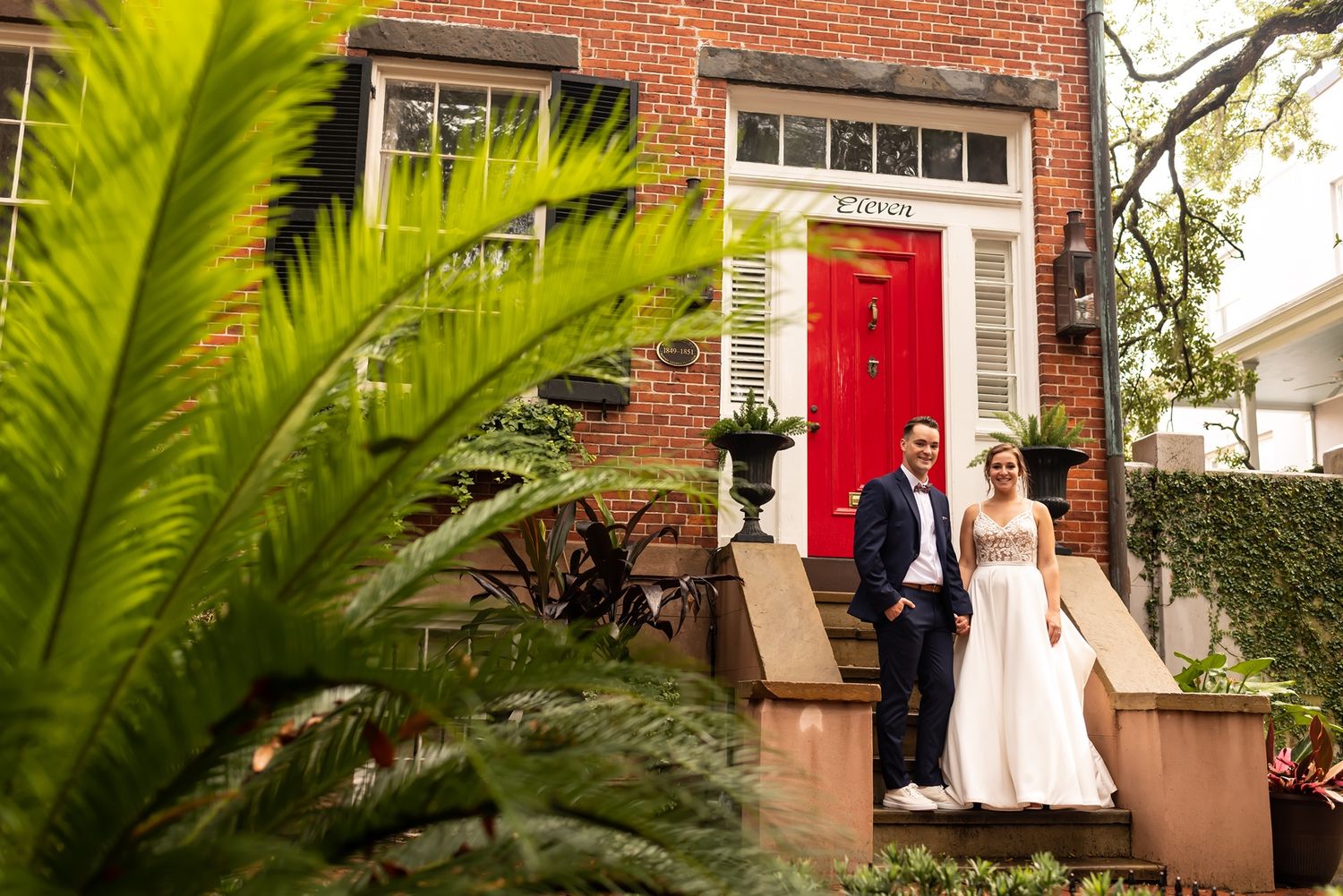 Bride and groom pose for photos on a brick pathway with lush greenery outside historic red-brick colonial building.