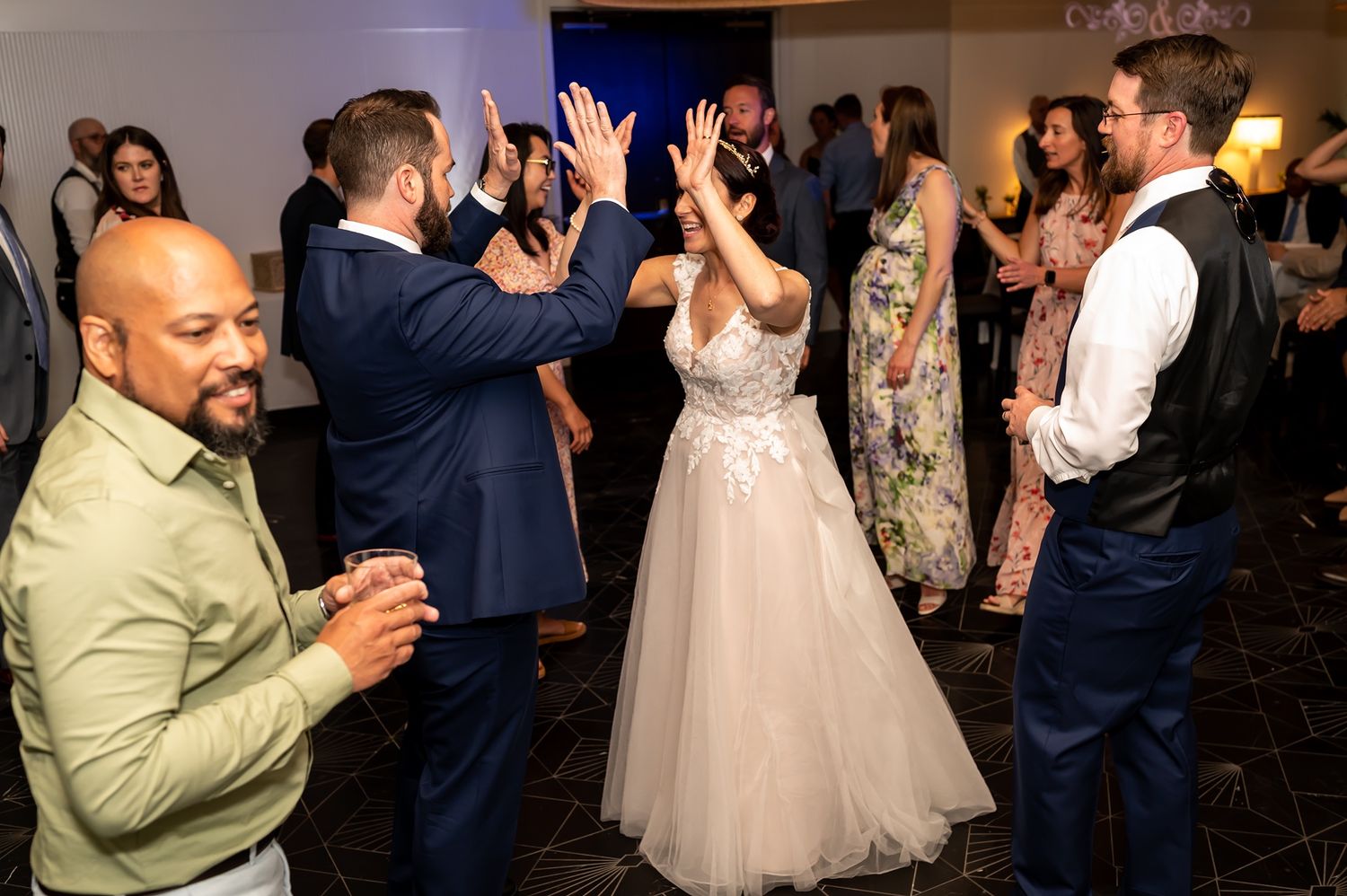 Wedding guests on a dance floor celebrate with the couple during a reception while a guest in a light green jacket looks on.
