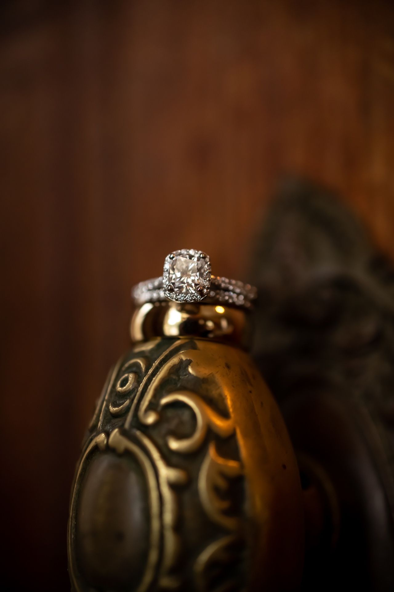 Diamond engagement ring with pave band rests on ornate metallic surface with vintage scrollwork design in warm lighting.